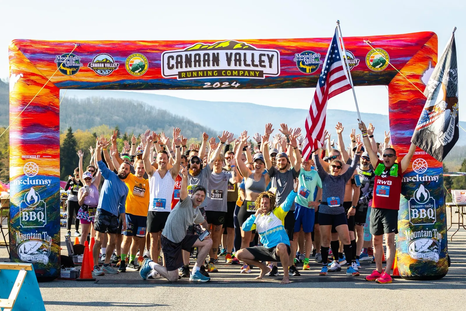 A large group of runners cheer and raise their hands under a colorful inflatable arch at the start line of the Canaan Valley Running Company 2024 race, with an American flag waving.