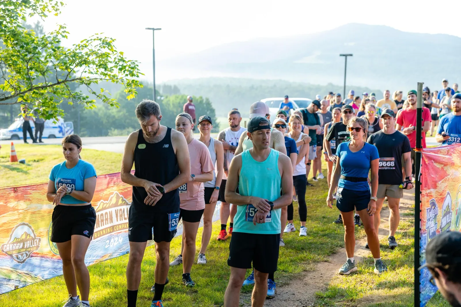 A group of runners in athletic wear stand in a line, some looking down at their watches, others smiling, with a scenic mountain landscape in the background and a colorful race banner in the foreground.