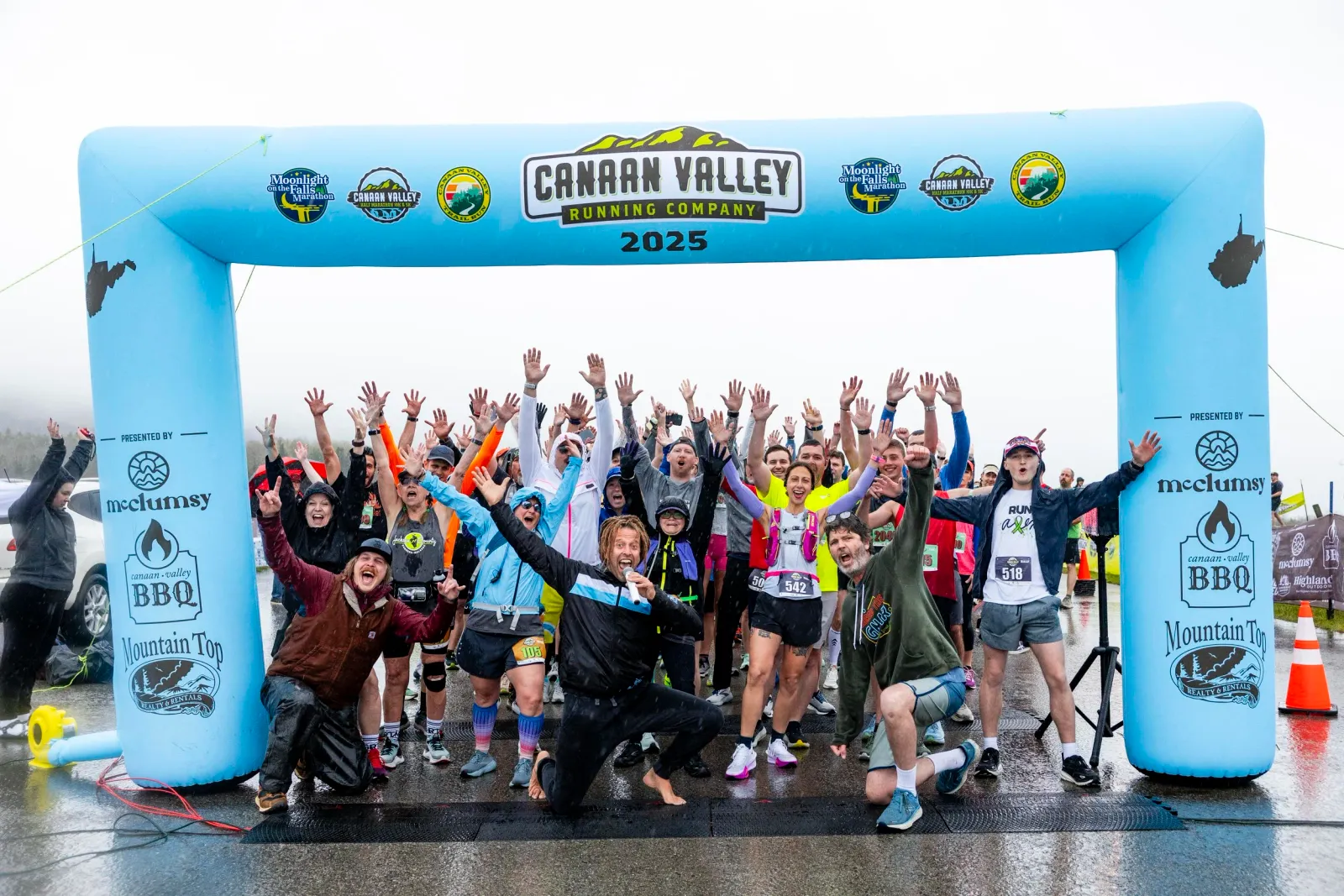 A large group of runners cheer and raise their hands under a blue inflatable arch at the start of the Moonlight on the Falls Marathon.
