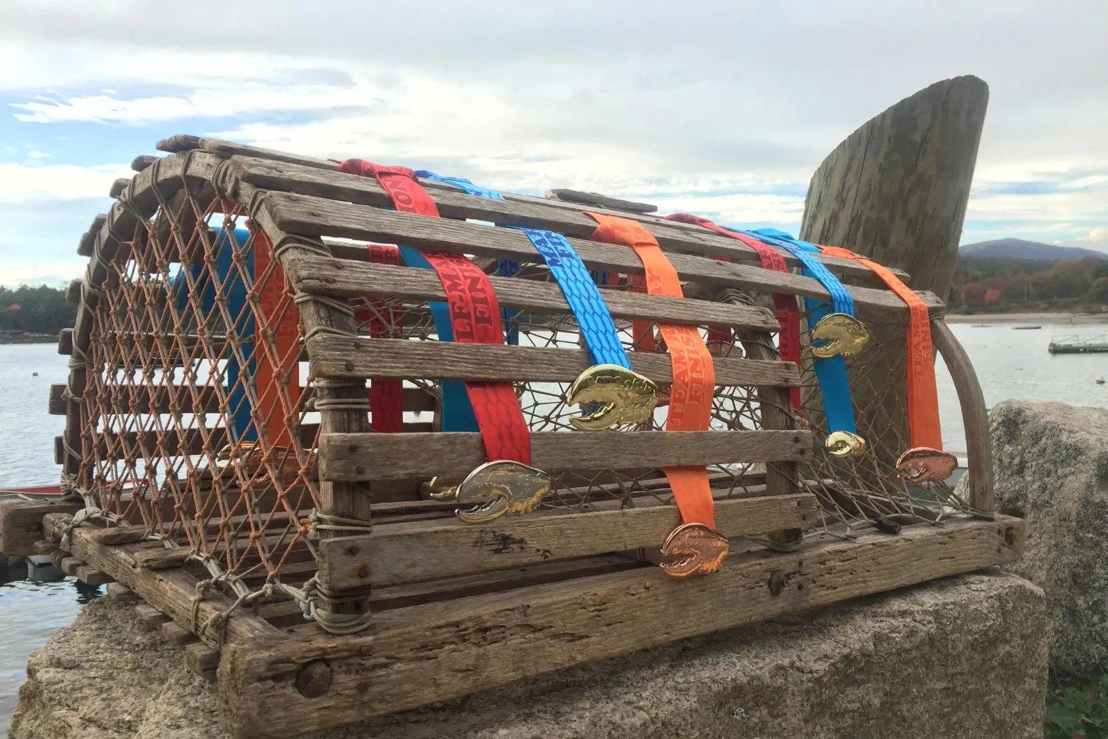 Race medals with lobster-shaped charms hanging from a wooden lobster trap near the water.