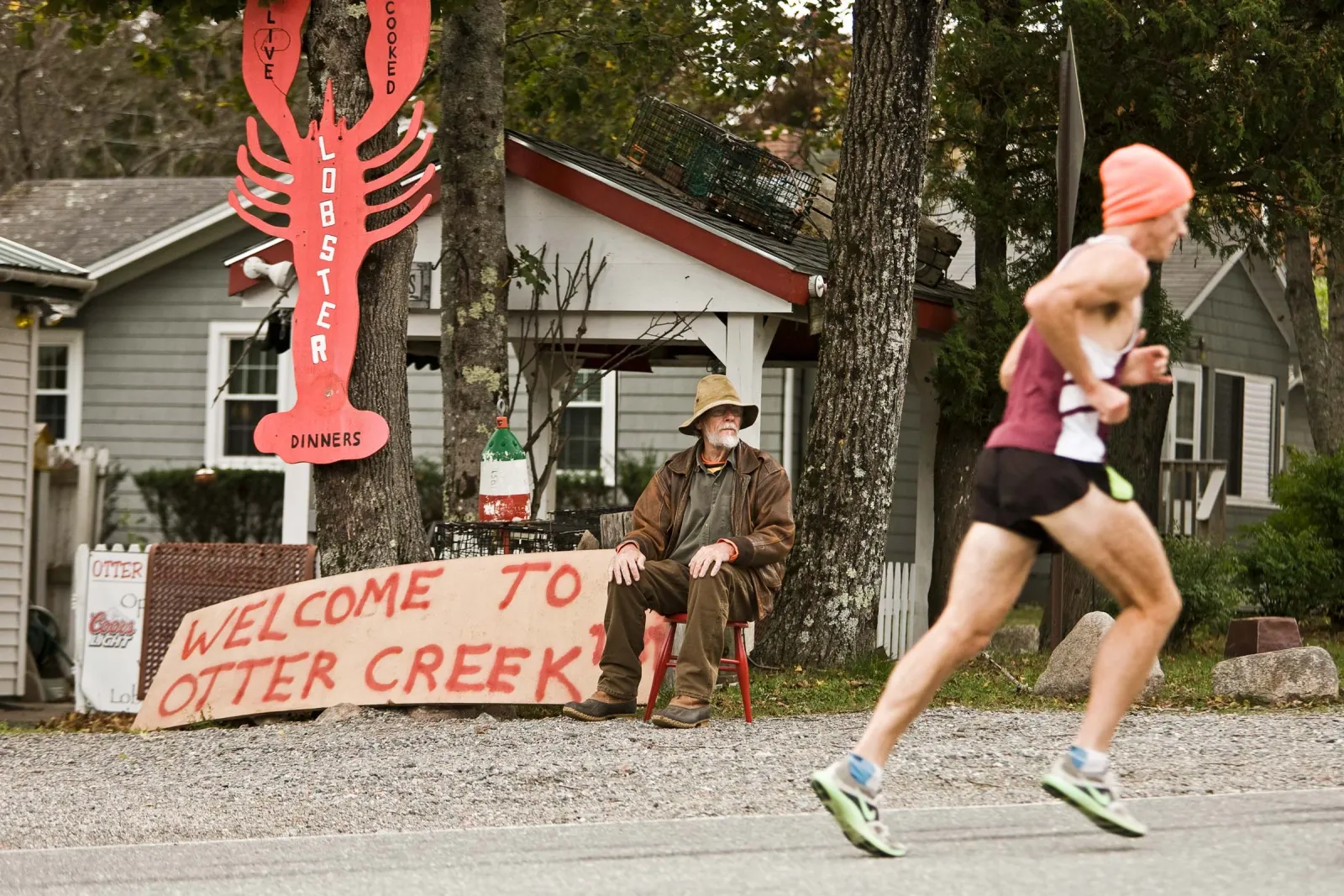 A runner in an orange beanie and a maroon and white singlet runs past a man sitting on a red stool next to a large red lobster sign and a 'Welcome to Otter Creek' sign.