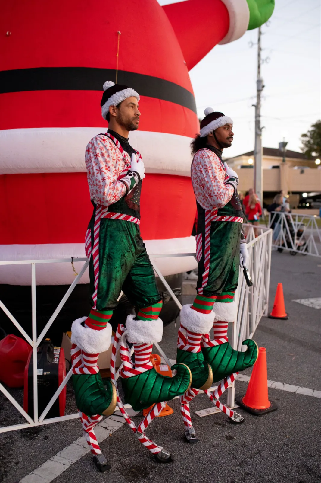 Two people dressed as elves on stilts stand in front of a large inflatable Santa Claus decoration.