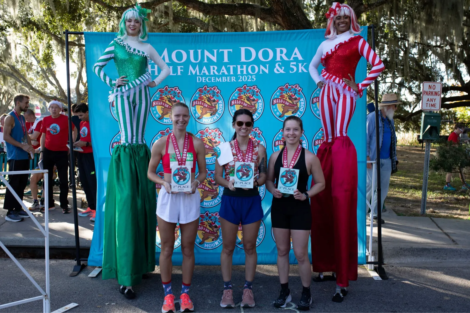 Three female runners, winners of the Mount Dora Half Marathon & 5K, pose with their medals in front of a race banner and two costumed performers.