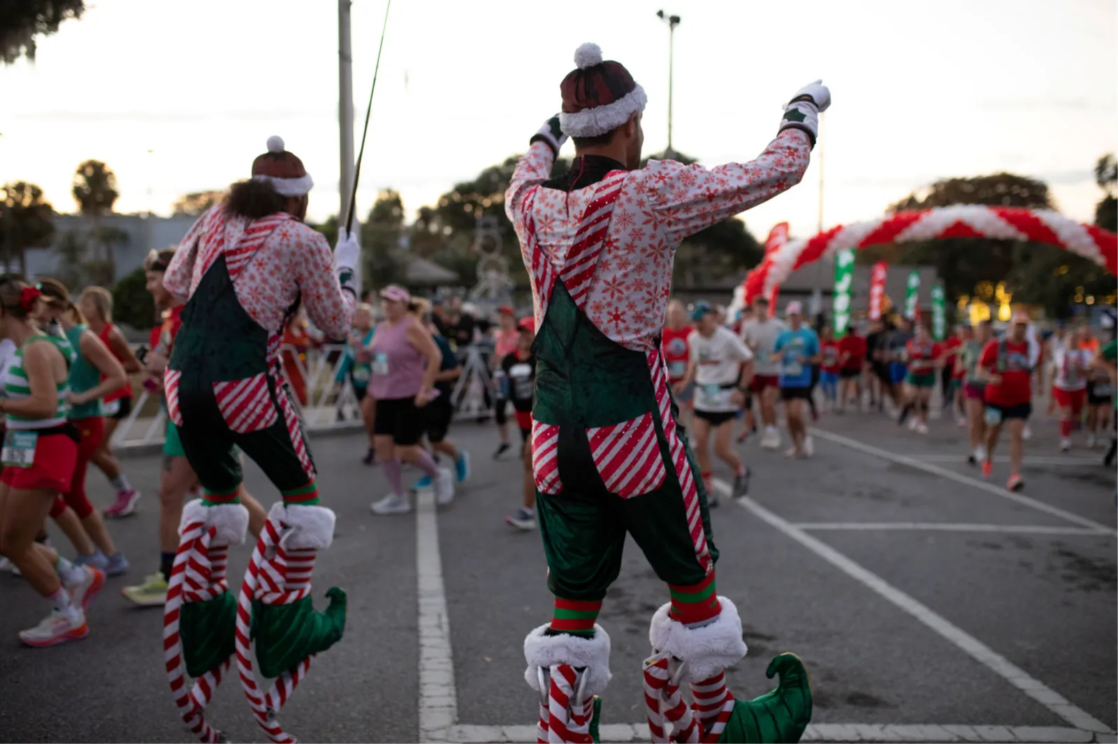 Runners in festive elf costumes on stilts at the start of a race, with other participants in the background.