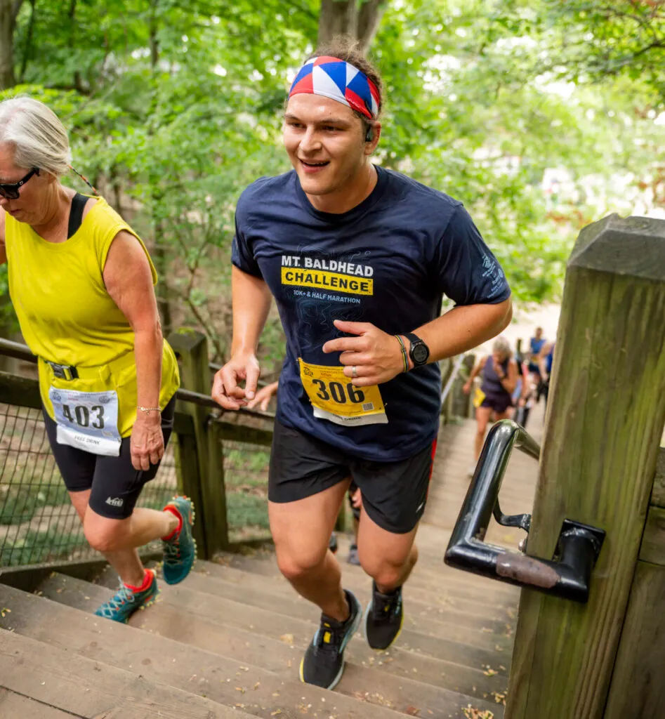 A male runner wearing a navy blue Mt. Baldhead Challenge t-shirt and a colorful headband runs up a wooden staircase during a race, with a female runner in a yellow shirt ahead of him.