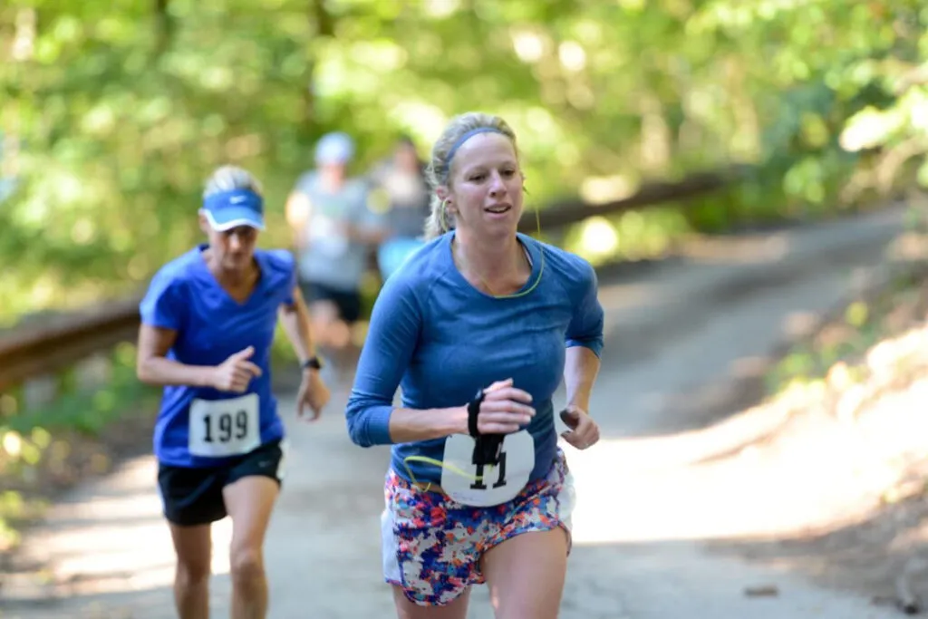 Two female runners on a dirt path surrounded by trees, one in the foreground wearing a blue long-sleeved shirt and colorful shorts, the other behind her in a blue t-shirt and black shorts.