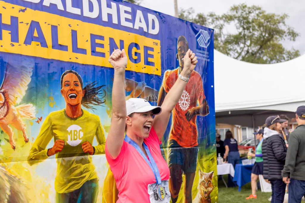 A woman with her arms raised in celebration, wearing a pink shirt and a race medal, stands in front of a large banner for the 'Baldhead Challenge'.