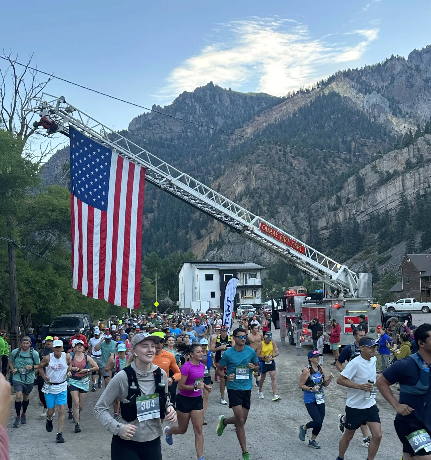 Runners starting a race with a large American flag displayed from a fire truck ladder, with mountains in the background.