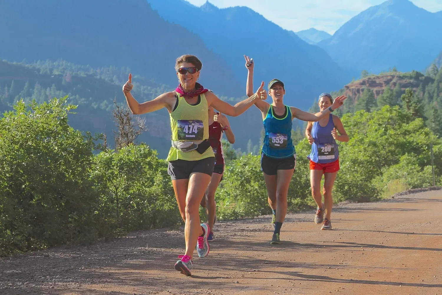 Runners smiling and giving thumbs up while running on a dirt road with scenic mountains in the background.
