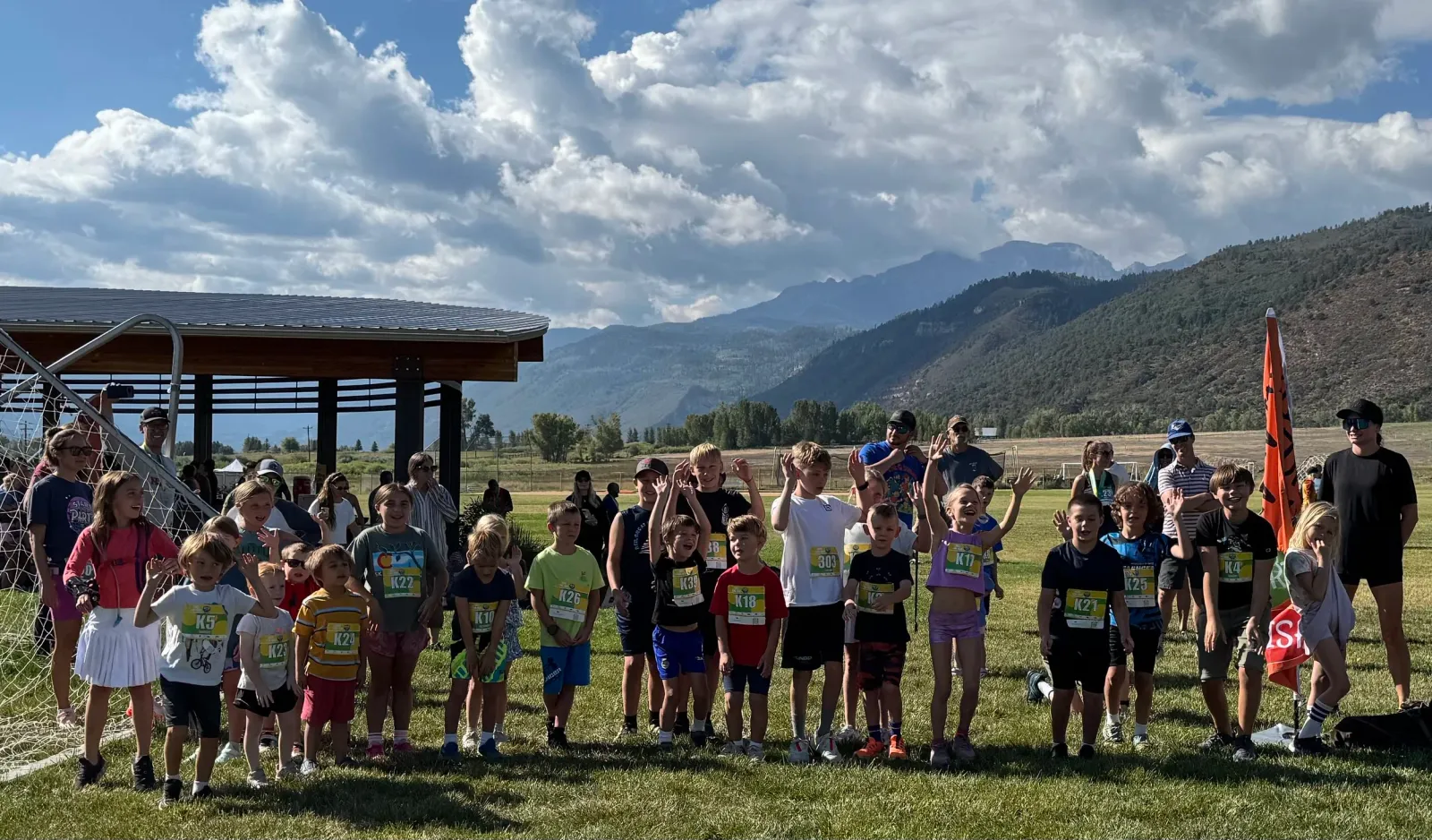A group of children wearing race bibs stand on a grassy field, with mountains in the background, smiling and waving.