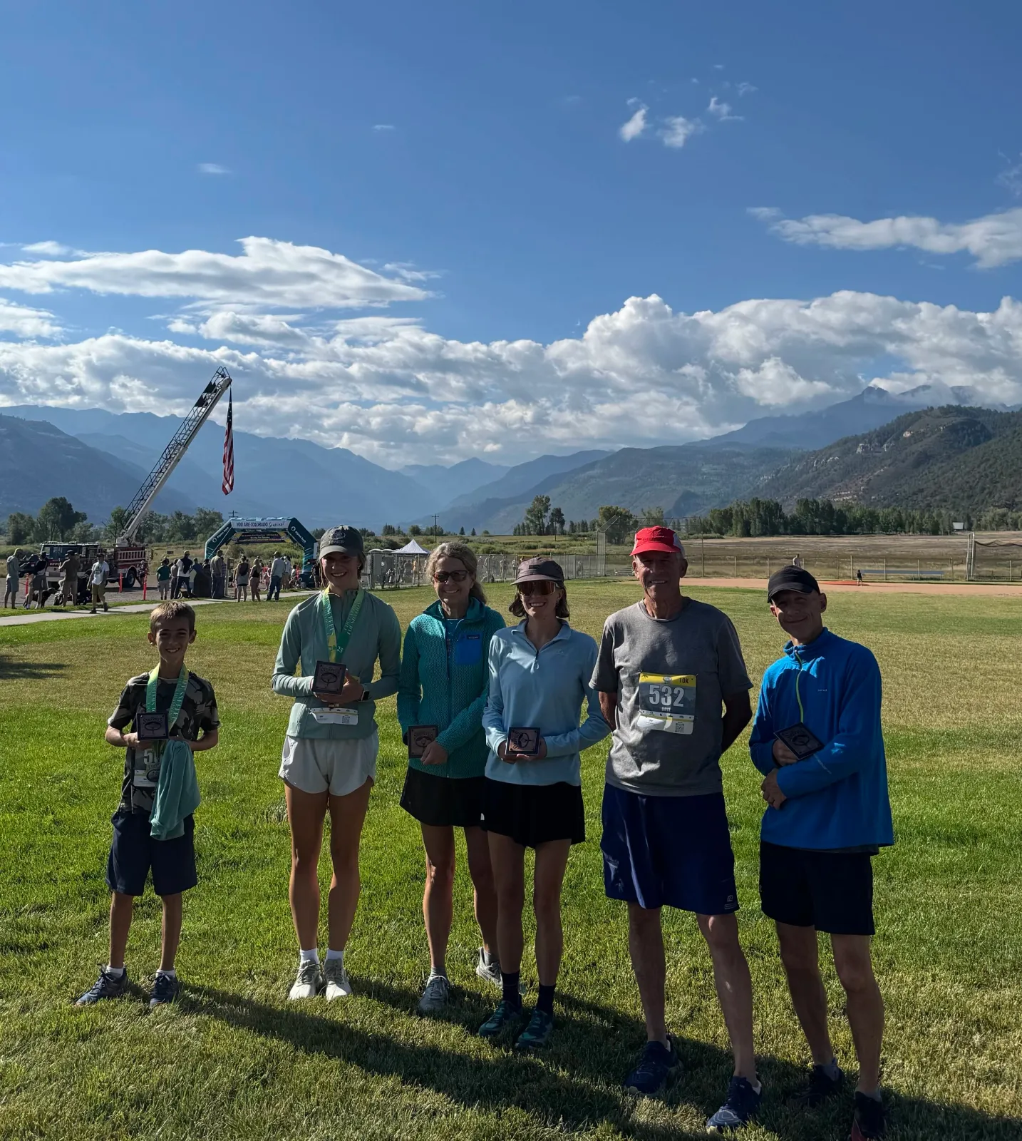 Five race winners, including a young boy and four adults, pose for a photo holding their medals with a scenic mountain range in the background.