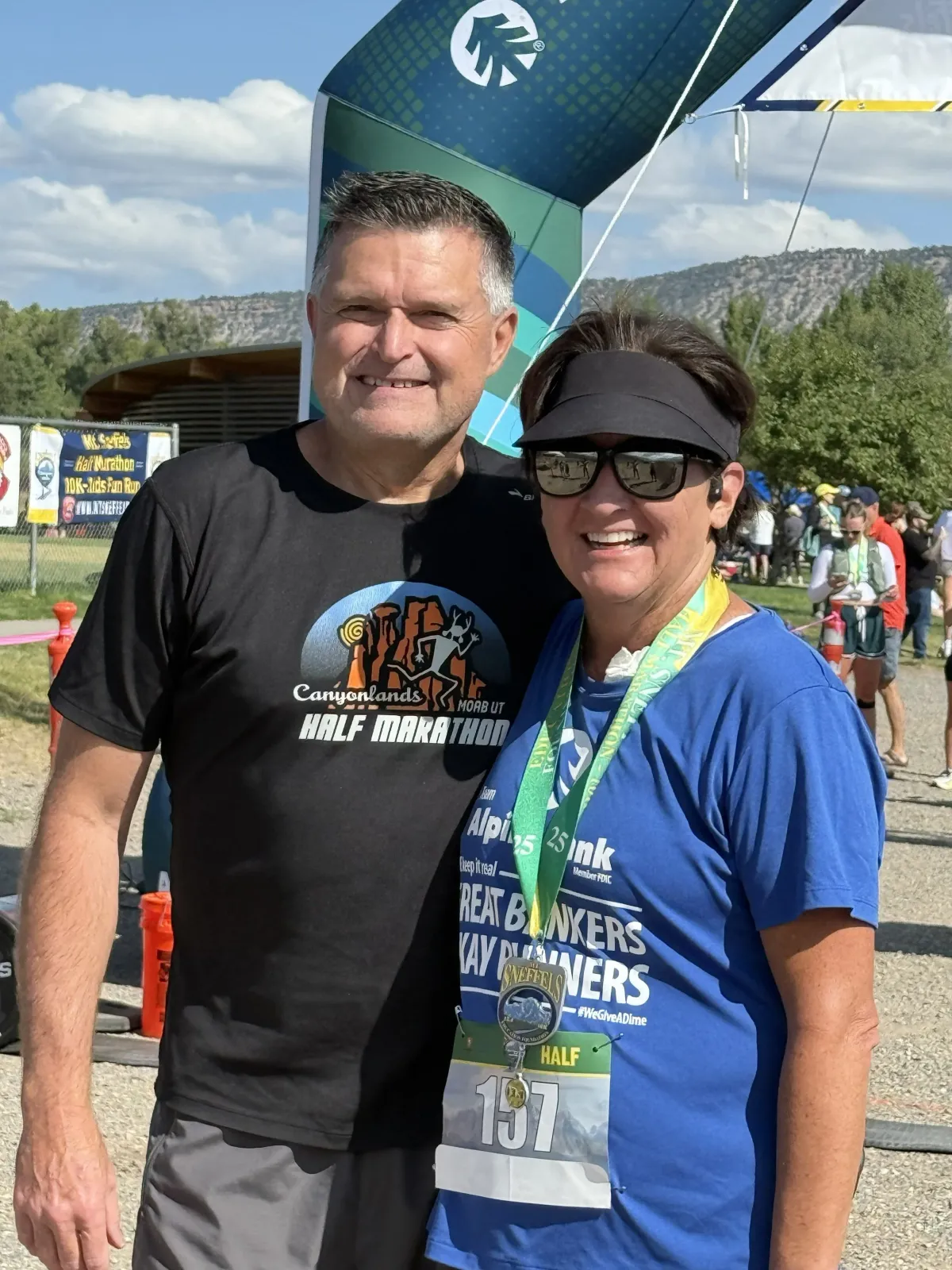 A man and a woman smiling at the camera, both wearing race medals, with a finish line arch in the background.