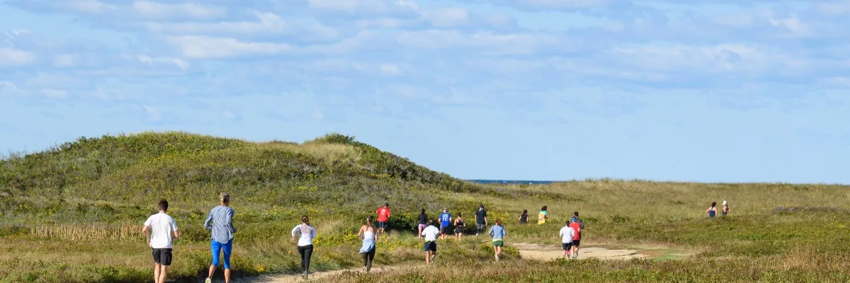 Runners on a path through grassy dunes with the ocean in the distance.