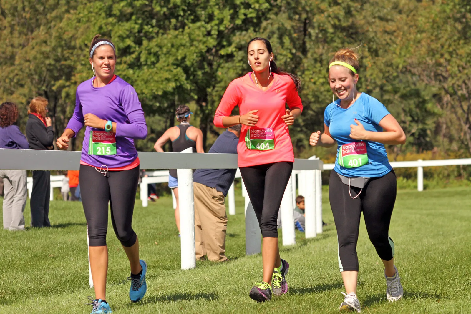 Three female runners in colorful shirts and black leggings running on a grassy course, with race bibs visible.