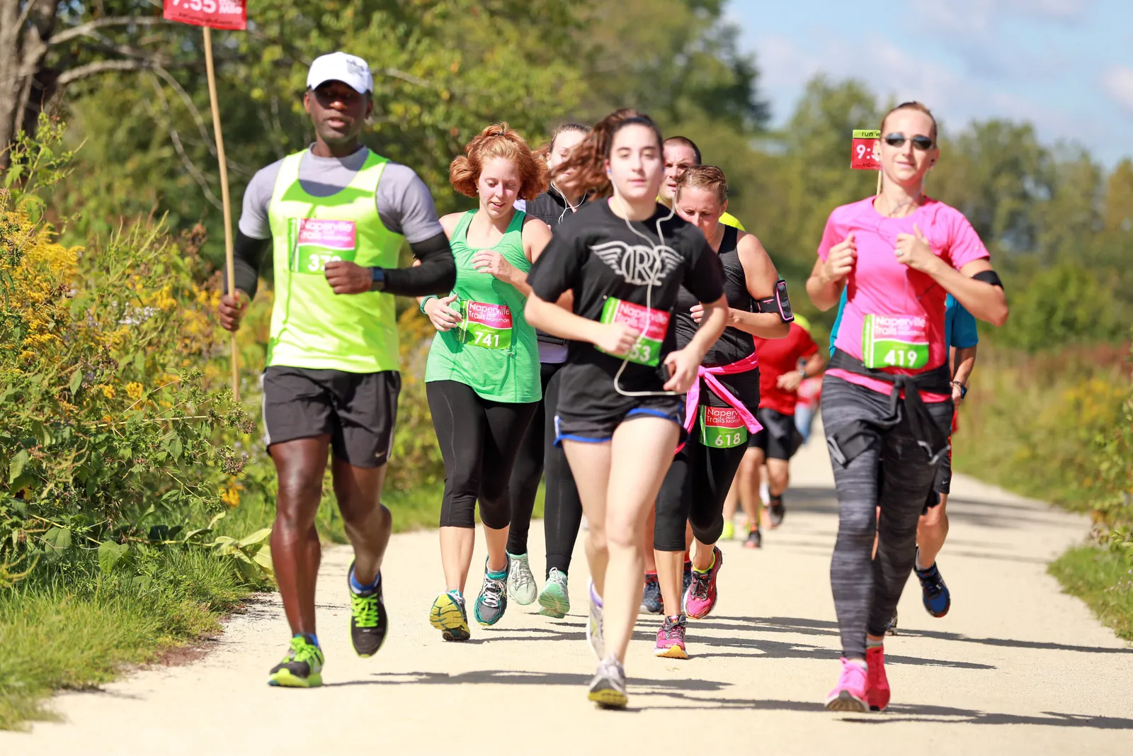 A group of runners on a dirt path, with trees and foliage on either side. The runners are wearing race bibs with 'Naperville Trails Marathon' visible.