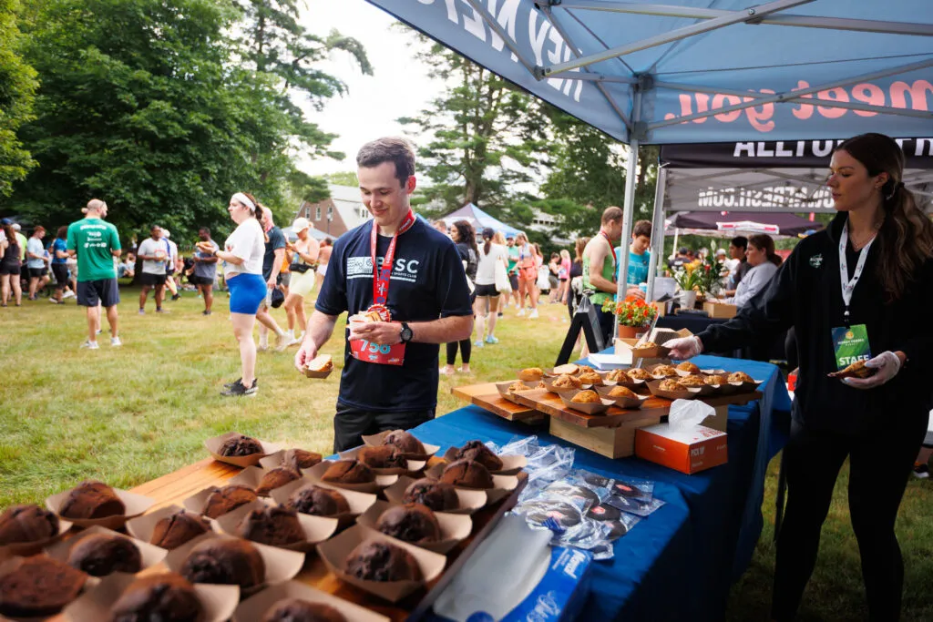 A runner in a black t-shirt with a bib number smiles as he holds a muffin, with other runners and tents in the background.