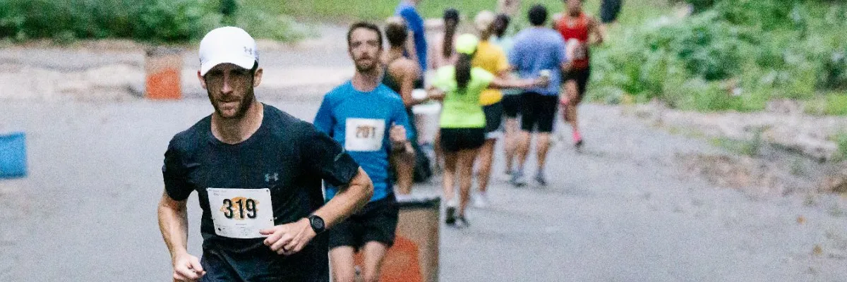 A male runner wearing a white cap and black t-shirt with bib number 319, looking towards the camera while running on a paved road with other runners in the background.
