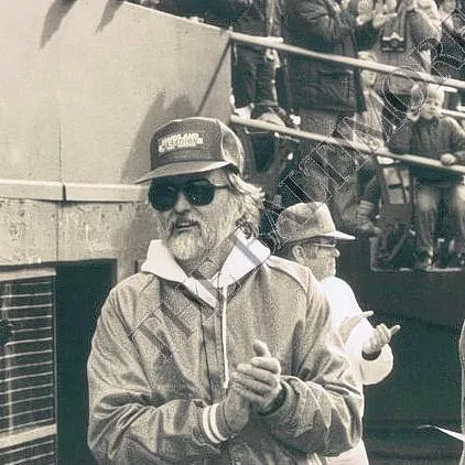 A man with a beard and sunglasses, wearing a baseball cap and jacket, claps his hands while standing in front of a crowd at a race event.