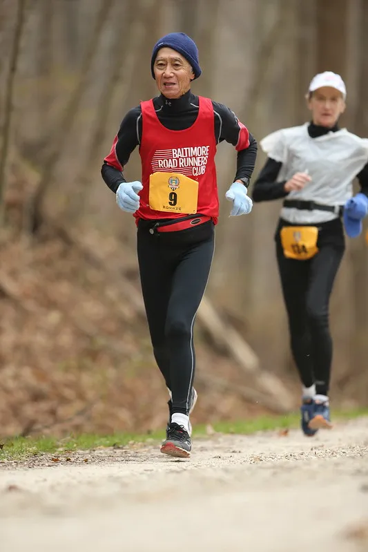 A male runner wearing a red Baltimore Road Runners Club singlet and a blue beanie runs on a dirt path, with another runner blurred in the background.