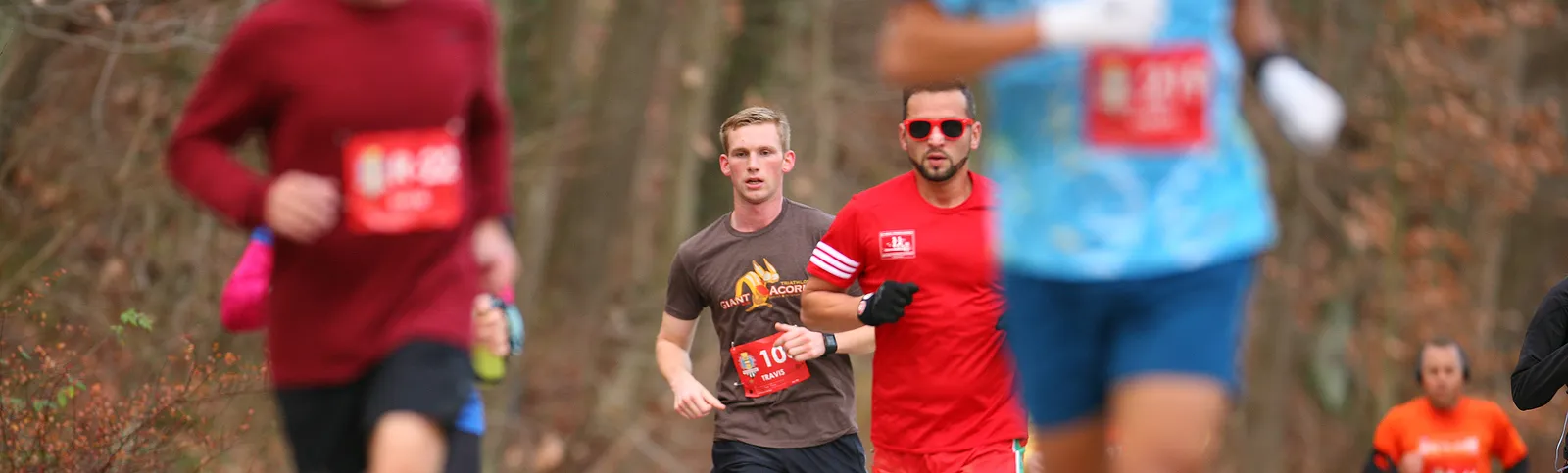 Runners on a wooded course, with one runner in the foreground wearing a brown shirt with a bib number 10.