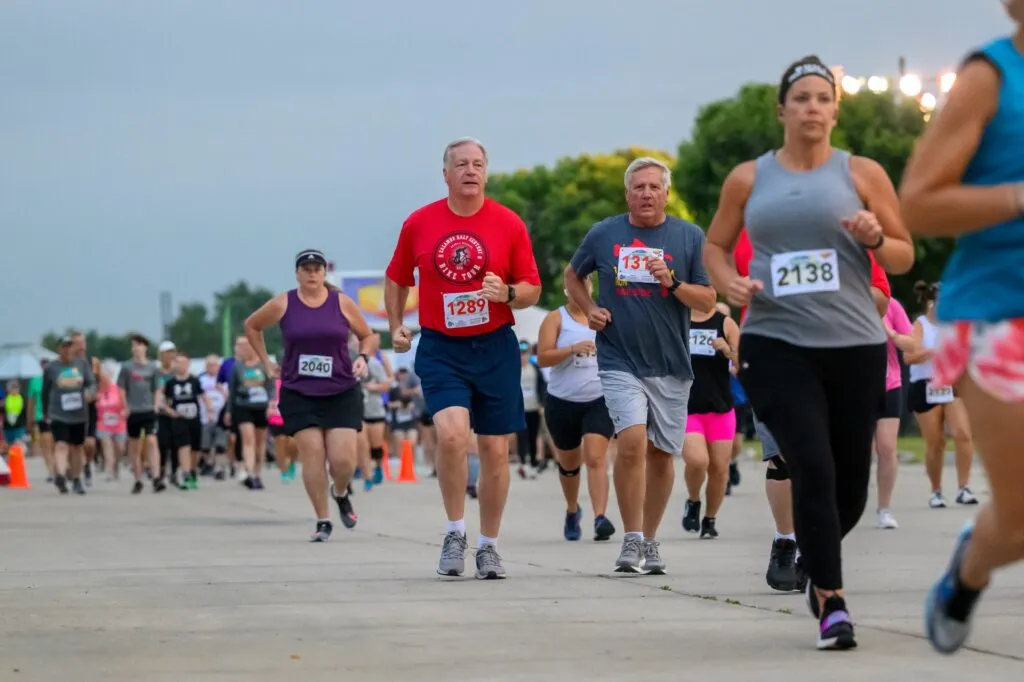 A group of runners in the foreground and background are running on a paved surface, with some wearing bib numbers.