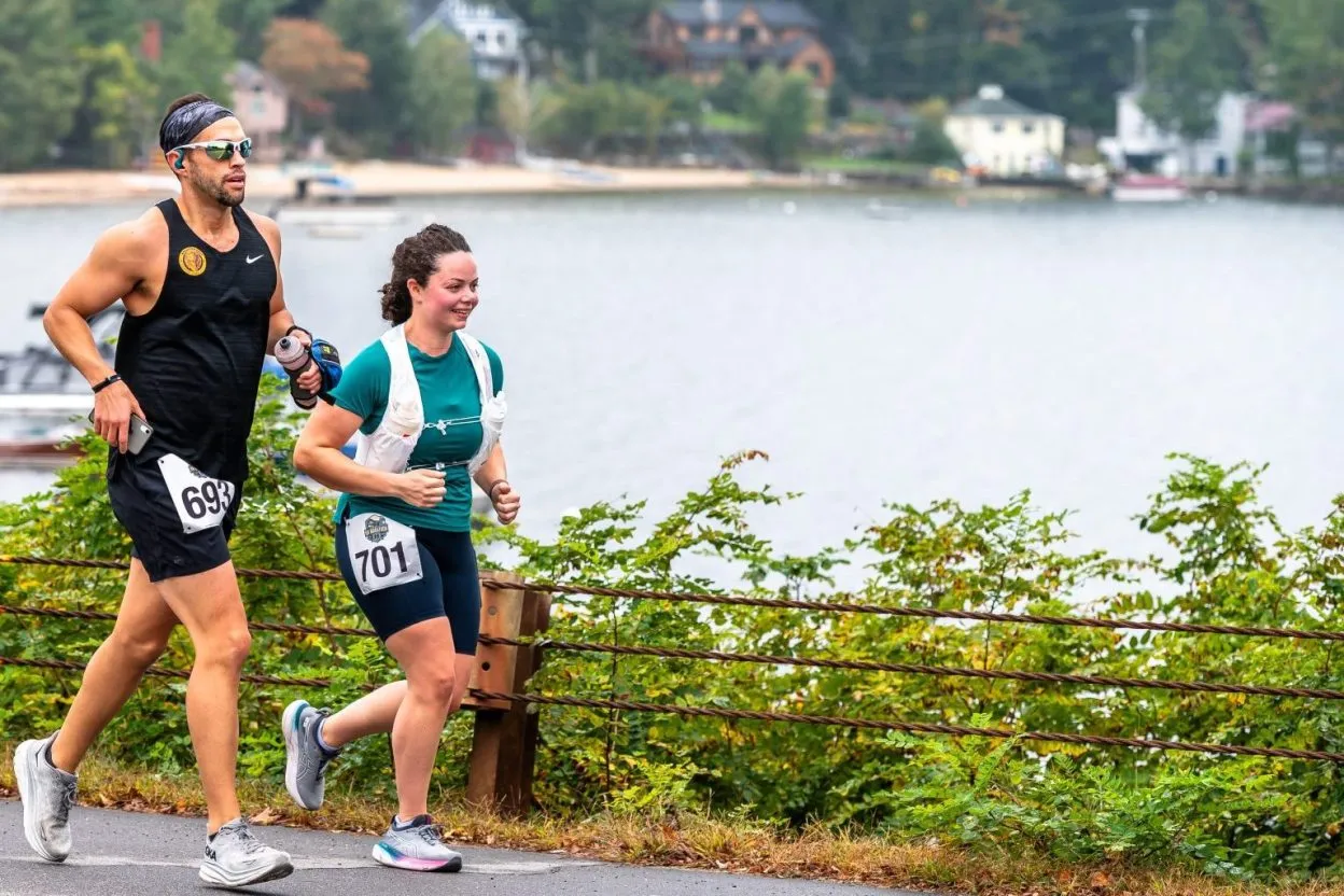 Two runners, a man and a woman, run along a road with a body of water and houses in the background.