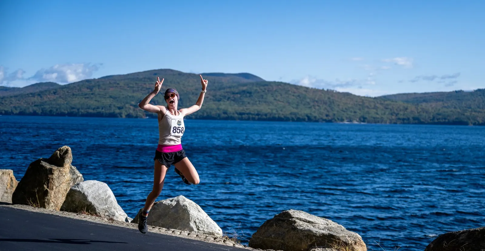 A runner in mid-air jump with arms raised in celebration, wearing bib number 858, with a large blue lake and tree-covered hills in the background.