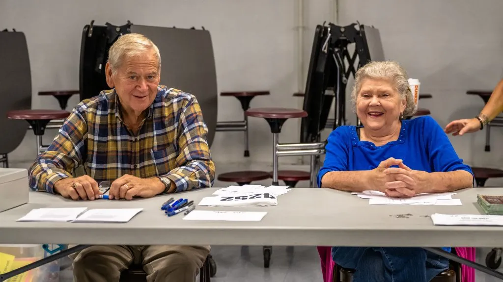 A man and a woman smiling while sitting at a registration table for a race.