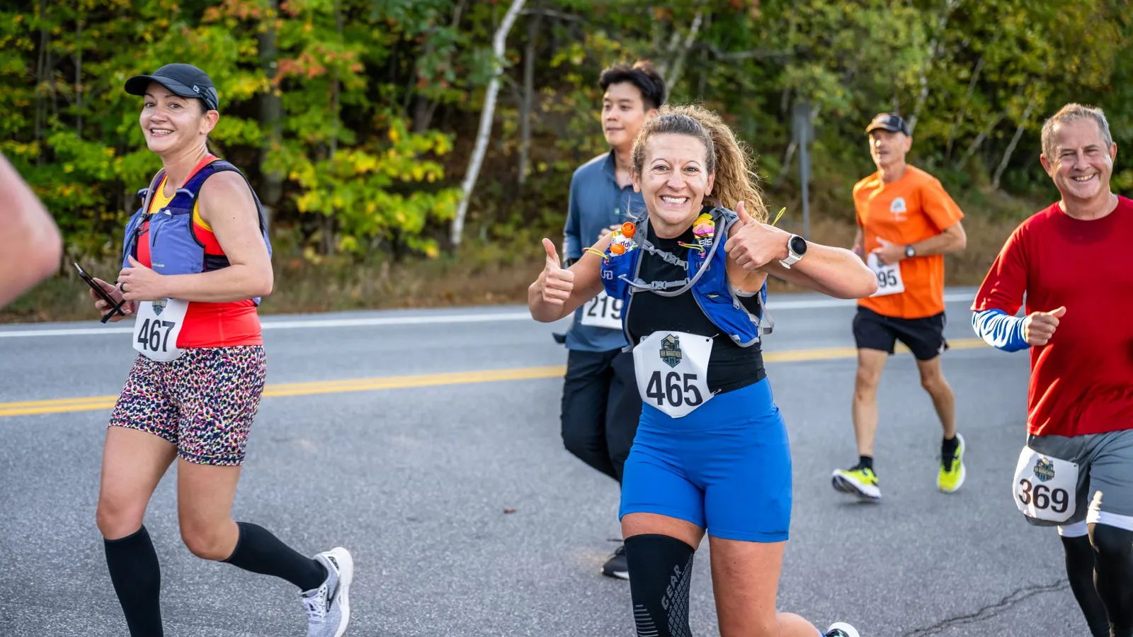 A female runner with bib number 465 smiles and gives two thumbs up while running on a road, with other runners in the background.