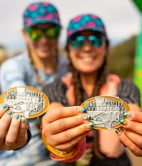 Two smiling runners in colorful hats and sunglasses hold up their Dinosaur Valley Endurance Run 100 mile medals.