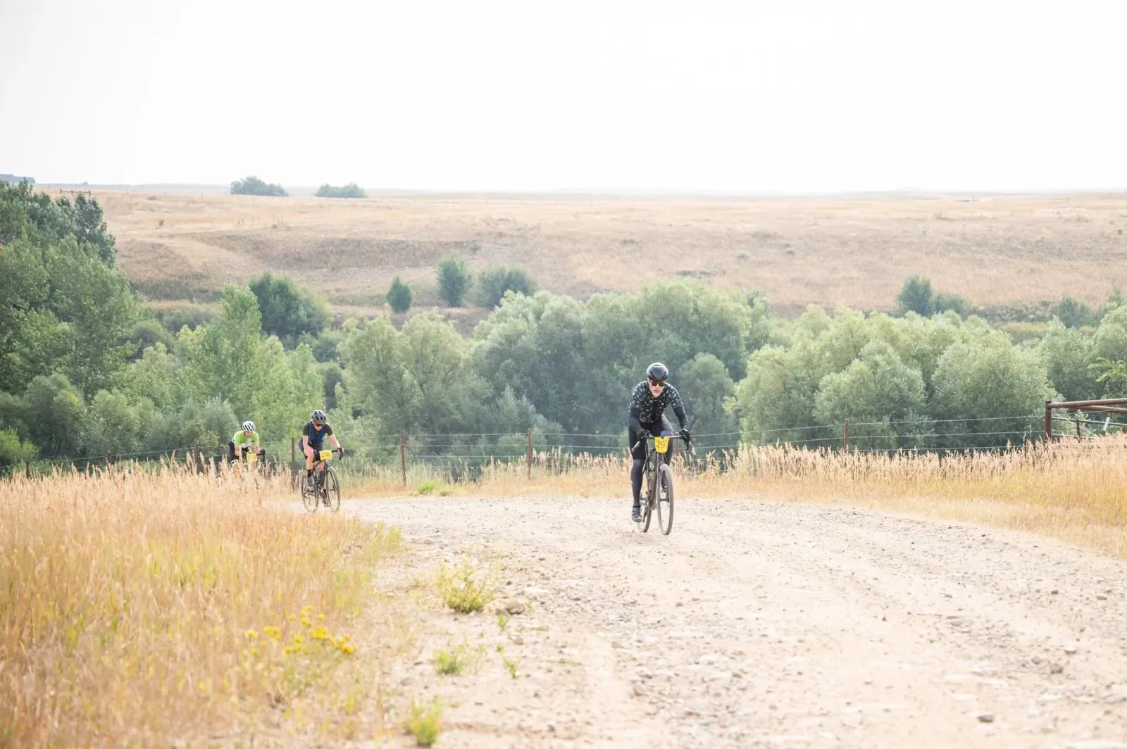 Cyclists riding on a dirt road through a dry, grassy landscape with trees in the background.
