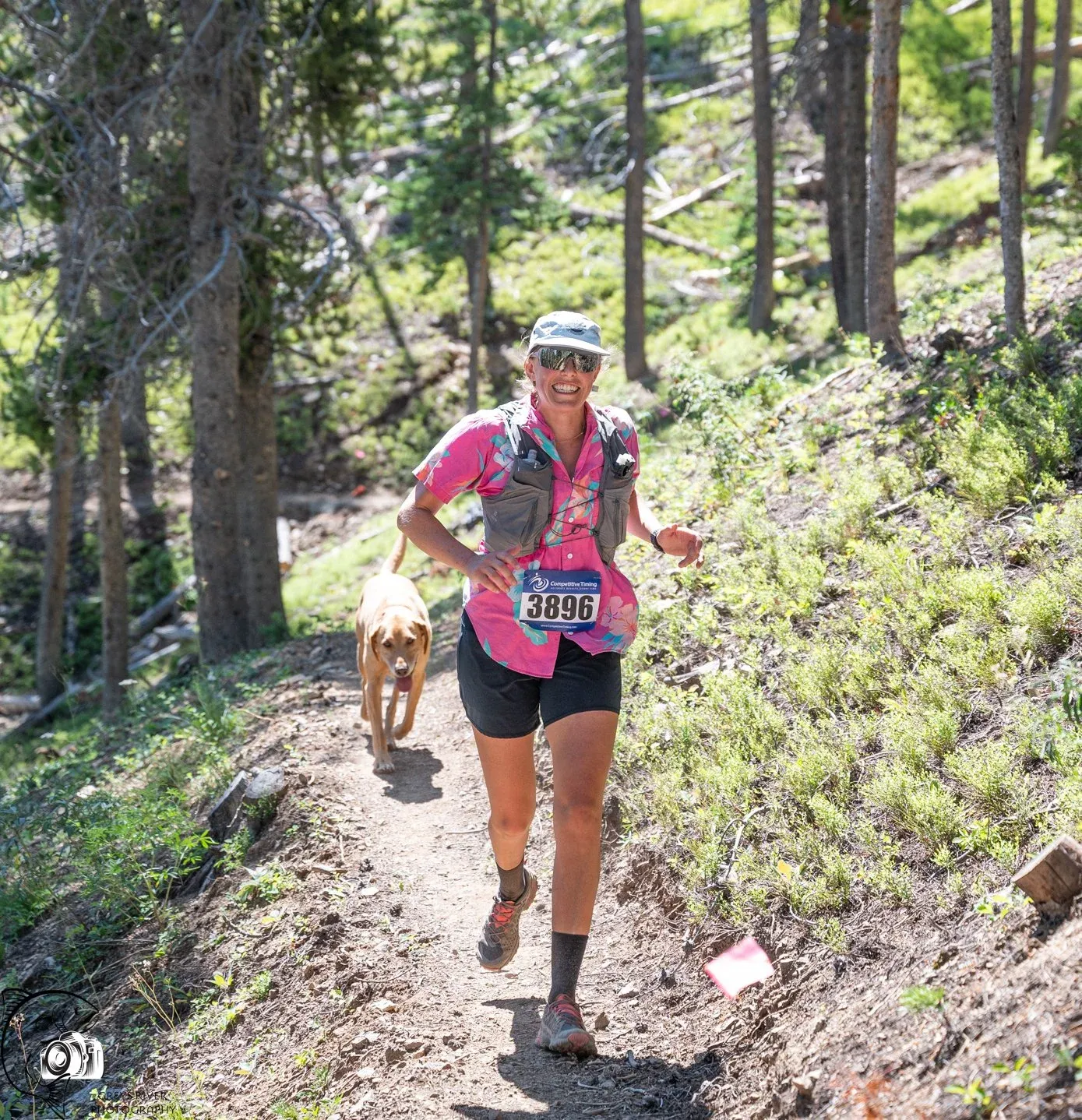 A female runner with a dog on a wooded trail during a race.