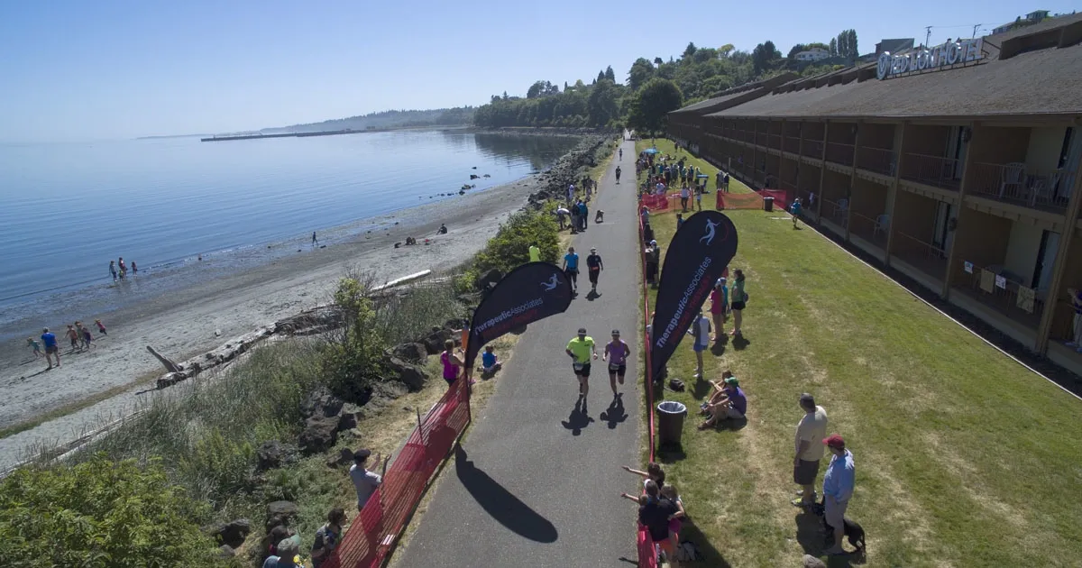 Runners on a paved path along a waterfront, with spectators and a hotel in the background.