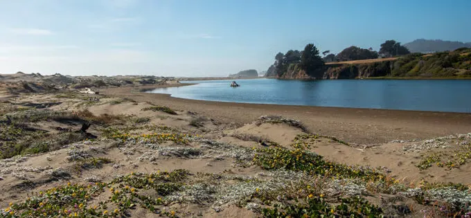 Scenic view of sand dunes and a body of water with trees and hills in the background.