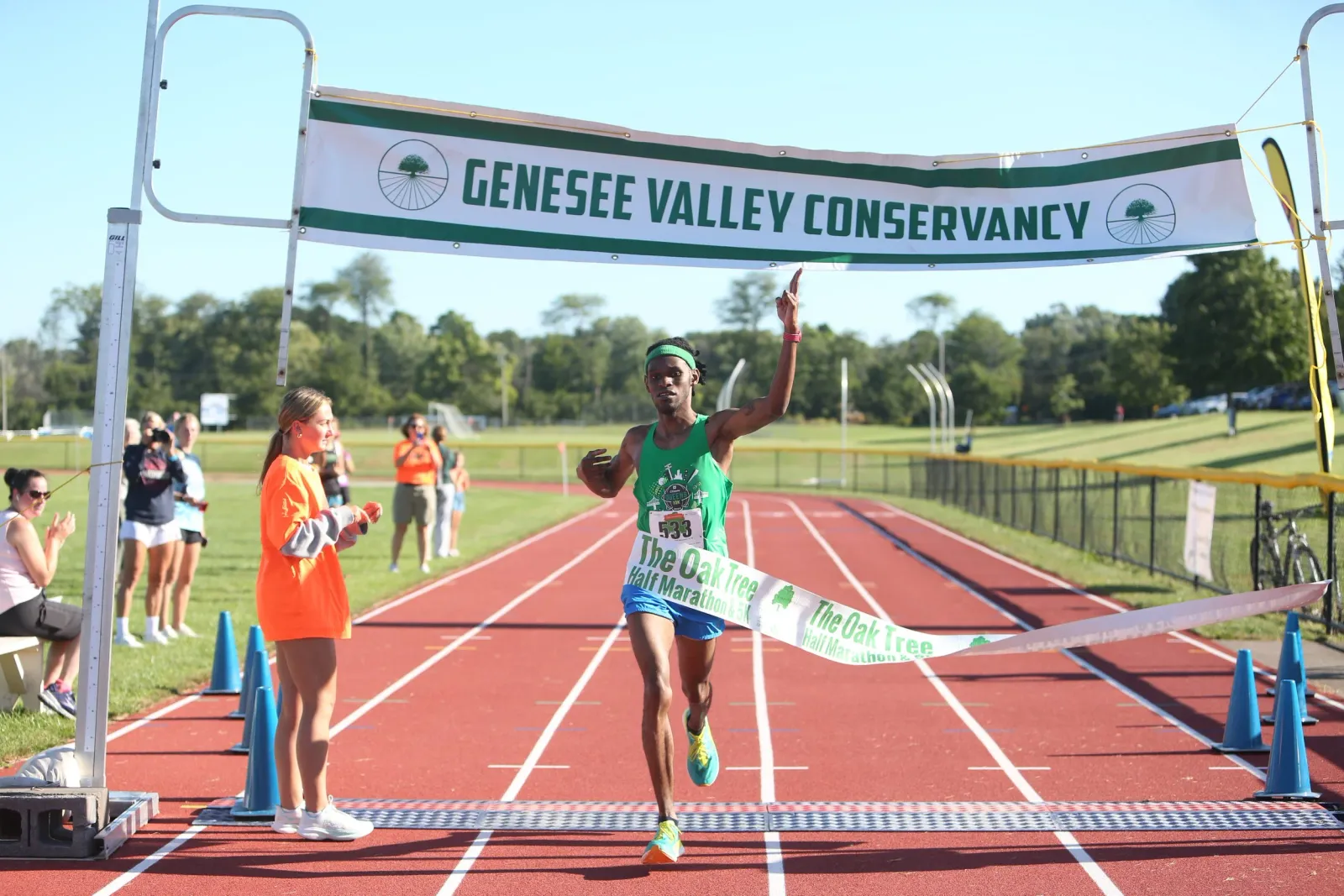 A male runner in a green singlet and blue shorts crosses the finish line, raising one arm in victory, with a banner overhead that reads 'Genesee Valley Conservancy'.