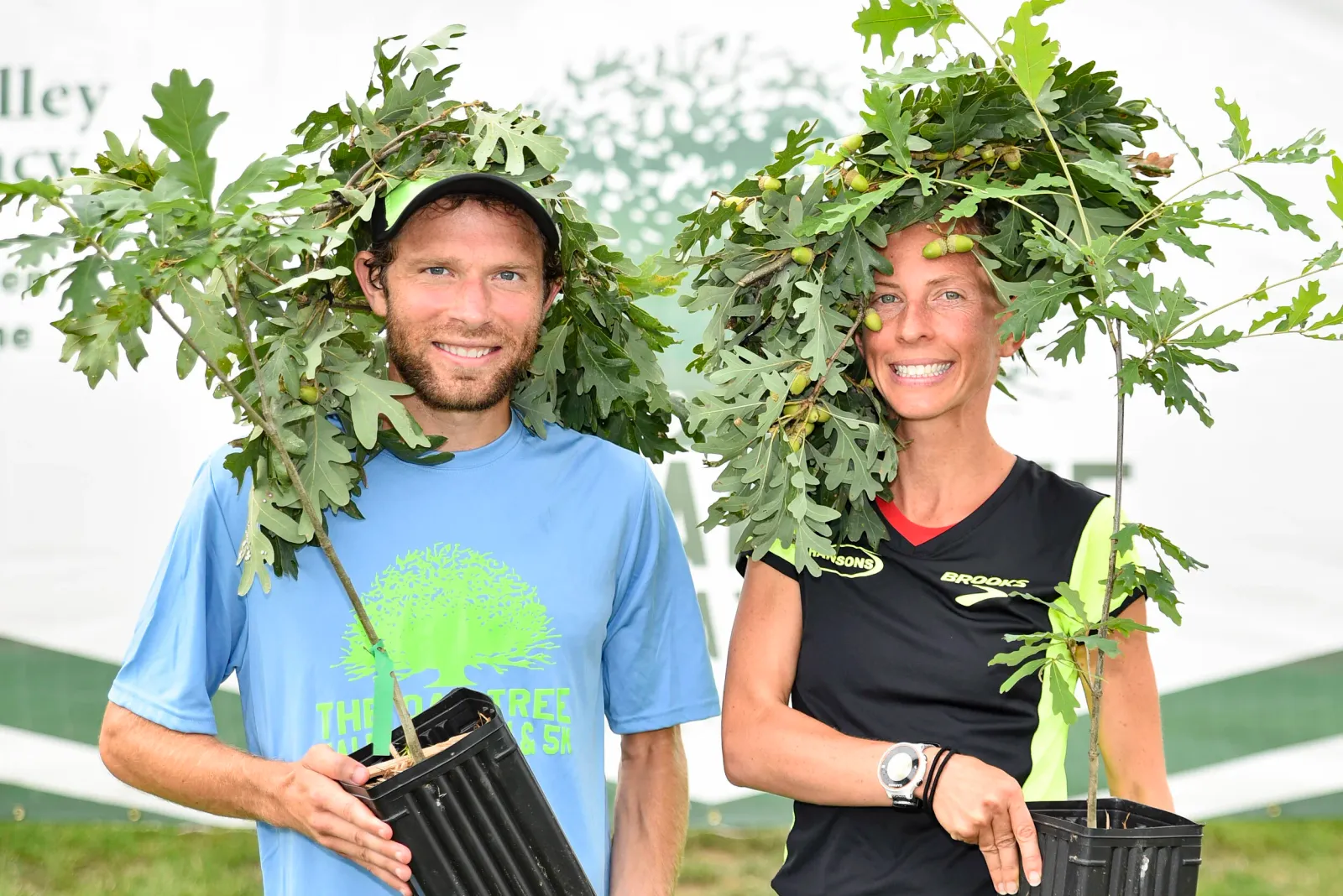 A man and a woman, both smiling and wearing oak branches with acorns on their heads, hold small oak saplings.