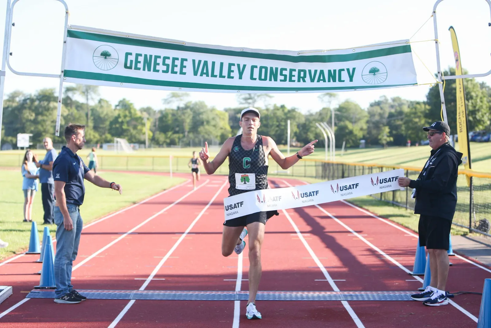 A male runner crosses the finish line, breaking a banner, with race officials and spectators in the background.
