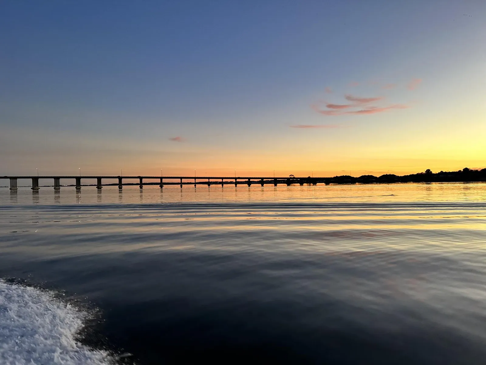 A long bridge stretches across calm water reflecting a colorful sunset sky.