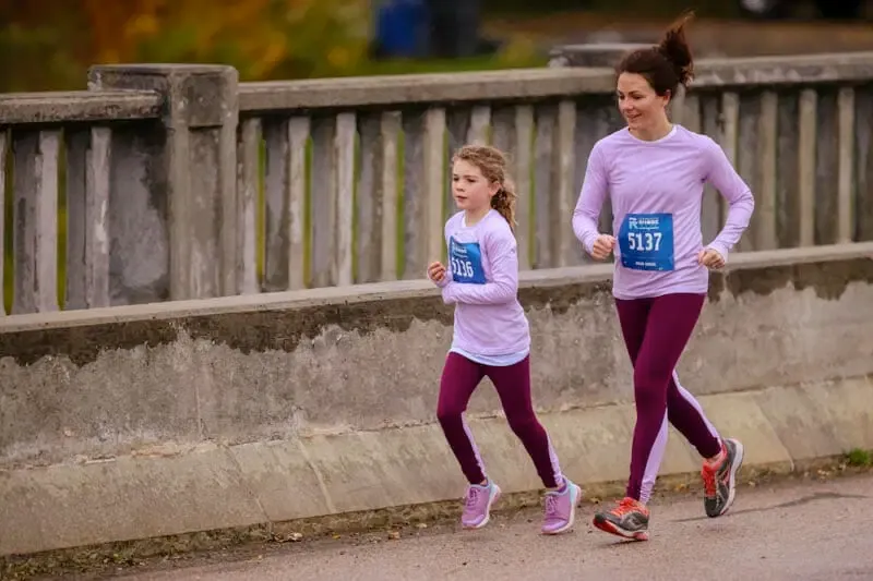 An adult and a child running a race, both wearing purple long-sleeved shirts and dark leggings, with race bibs visible.