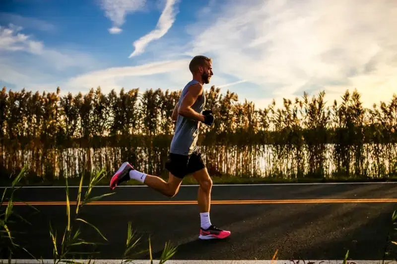 A male runner with a beard runs on a road with reeds and a cloudy sky in the background.