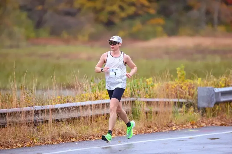 A male runner wearing a white tank top with bib number 3, black shorts, and bright green shoes runs on a wet road with dry grass and trees in the background.