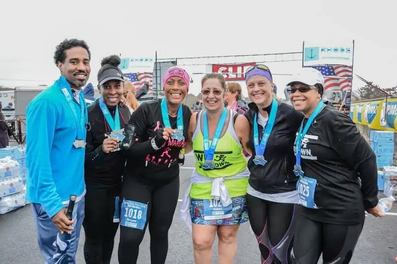 A group of six runners, all smiling and wearing medals, pose for a photo after completing a race. The finish line structure and American flags are visible in the background.