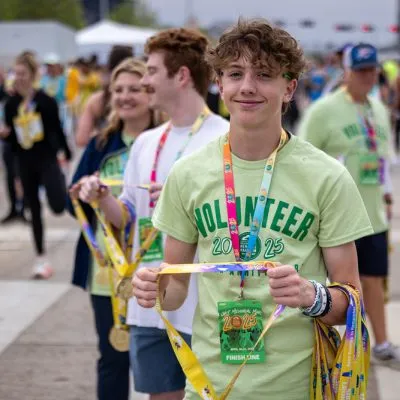 A young male volunteer smiles at the camera, holding several race medals and wearing a volunteer t-shirt.