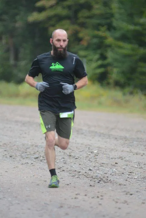 A male runner with a beard runs on a gravel path, wearing a black t-shirt and shorts.