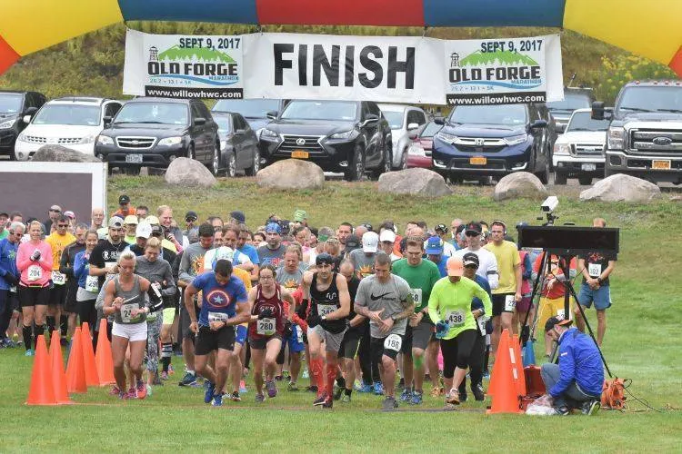 A large group of runners are gathered at the start line of the Old Forge Marathon, with a banner overhead that reads 'FINISH'.