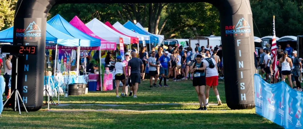 Runners gather near the finish line arch with tents and an American flag in the background.