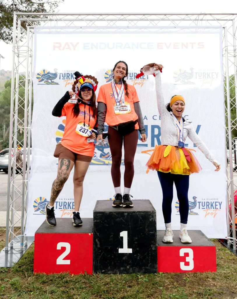 Three women on a podium celebrating after a race, two wearing turkey-themed hats and one with a festive tutu.