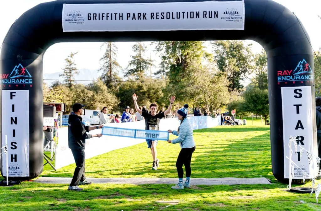 A runner crosses the finish line with arms raised in celebration, as two people hold the 'Overall Winner' banner at the Griffith Park Resolution Run.