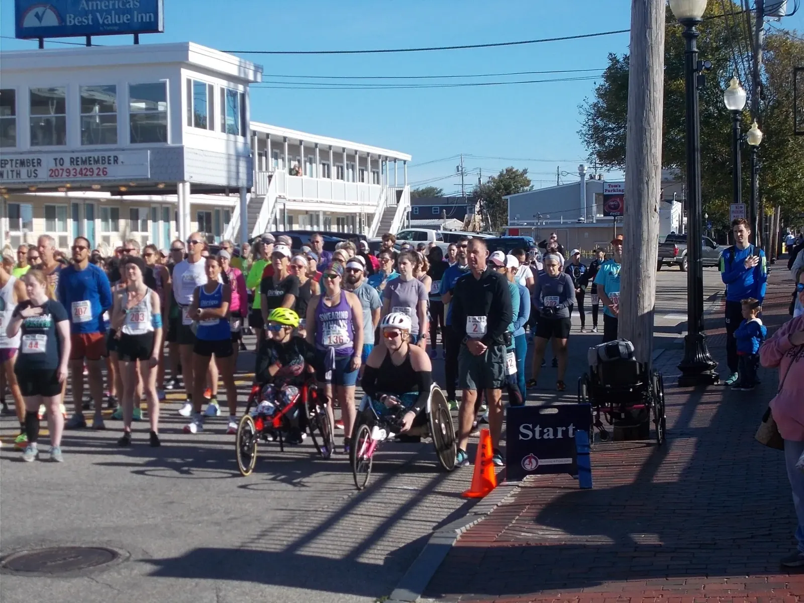 A group of runners, including some in racing wheelchairs, gathered at the start line of a race on a sunny day.
