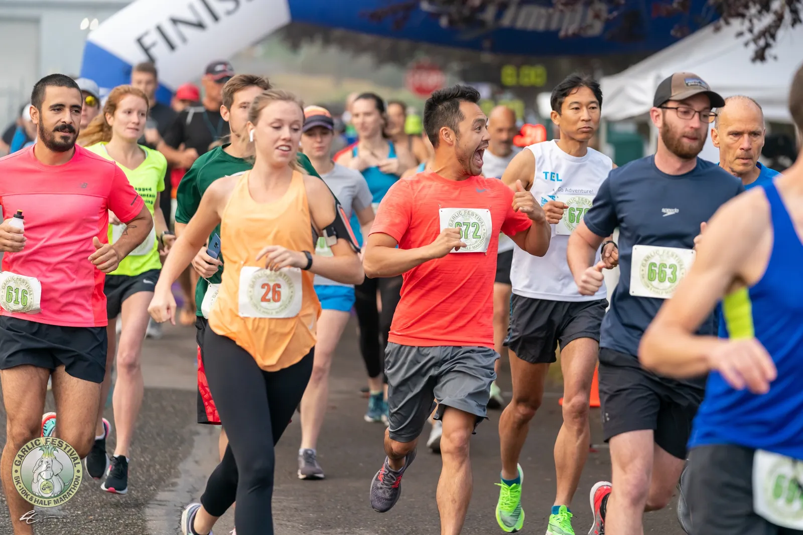 A group of runners in colorful shirts and bib numbers running towards the camera under a finish line arch.